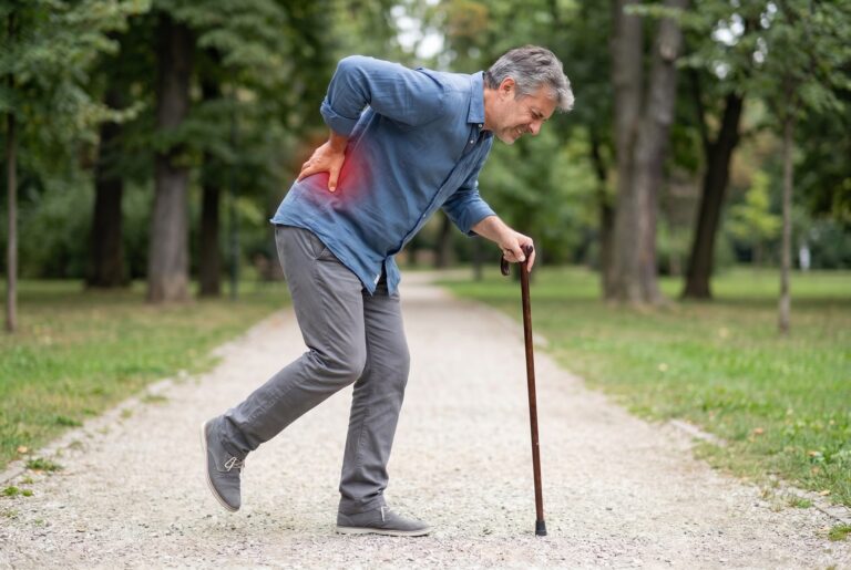 Older man walking with a cane on a park path and holding his lower back, a common sign of spinal stenosis