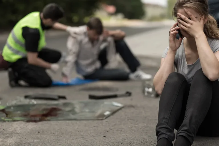 Woman sitting on the road on her phone after a car accident while a responder helps an injured man